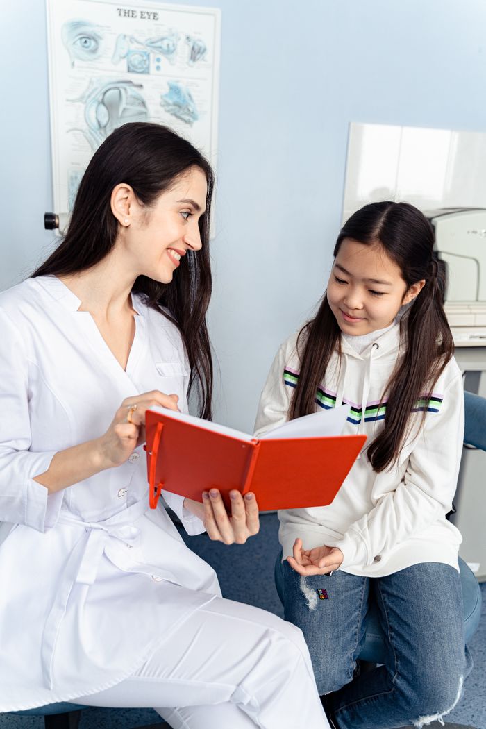 A female doctor shows a notebook to a young girl in a clinic setting, discussing eye anatomy.